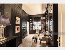 Elegantes Wohnzimmer mit dunklen Holzpaneelen, eingebauten Bücherregalen und Parkettboden im Fischgrätmuster. Der Raum ist möbliert mit einem Sofa, einem Polsterhocker, einem Lehnstuhl und einem Schreibtisch vor einem Fenster mit Blick ins Freie.