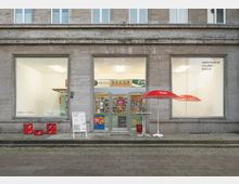 Außenansicht eines Gebäudes mit drei großen Schaufenstern; in der Mitte ein Eingang mit Glasdoppeltüren, flankiert von Fenstern mit der Aufschrift "Architektur Galerie Berlin". Vor dem Eingang stehen ein roter Sonnenschirm und einige Werbeelemente.