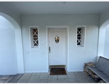 Helle Hauseingangspartie mit einer weißen Tür, flankiert von zwei kleinen Fenstern mit kreuzförmigen Gittern. Im Vordergrund helle Steinfliesen, rechts ein Holzstapel und eine schlichte Sitzbank.