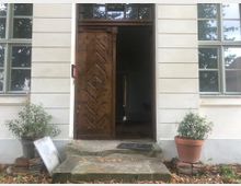 Ansicht eines Eingangsbereichs mit einer geöffneten Holz-Eingangstür in geometrischem Muster, flankiert von Blumen in Töpfen. Das Gebäude hat eine helle Fassade mit großen Fenstern und Stufen, die zum Eingang führen.