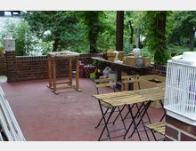 Teilweise überdachte Terrasse mit rotem Bodenbelag, umgeben von einer niedrigen Backsteinmauer. Im Vordergrund stehen Holztische und -stühle, daneben sind Kartons, Dekorationsobjekte und ein Holzgestell sichtbar. Im Hintergrund wachsen Bäume und Kletterpflanzen.