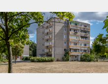 Wohnblock aus Beton mit fünf Etagen, mehreren Balkonen und gelben Akzenten an der Fassade, umgeben von Bäumen, Büschen und einer Rasenfläche. Im Hintergrund sind Wald und ein teils bewölkter Himmel sichtbar.