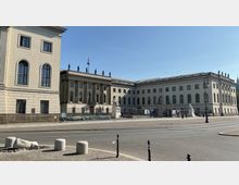 Straßenansicht eines historischen Gebäudekomplexes der Humboldt-Universität in Berlin, mit klassizistischer Fassade, Säulen und Statuen auf dem Dach. Im Vordergrund eine breite Straße, Bürgersteig und einzelne Poller.