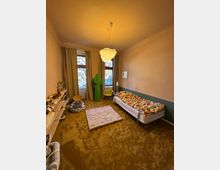 Ein schlicht eingerichtetes Zimmer mit einem Bett auf der rechten Seite, das mit einer bunten Decke bedeckt ist, sowie zwei Fenstern mit beigefarbenen Vorhängen im Hintergrund. Im Raum befinden sich ein Teppich, ein Regal an der linken Wand, eine grüne dekorative Kaktus-Skulptur und eine helle Hängelampe an der Decke.