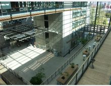 Großer, moderner Innenhof eines Gebäudes mit hohen Glasfassaden und Betonwänden. Der Bereich umfasst offene Sitzgelegenheiten, Pflanzen und viel natürliches Licht durch eine gläserne Dachkonstruktion.