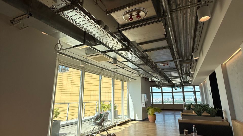 Modern office lounge corridor with light wood flooring, large floor-to-ceiling windows along one side, and an exposed industrial ceiling with visible ductwork, pipes, and cable trays. Low seating and planters line the right side, with a balcony railing and city view visible through the windows.