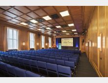 Konferenzsaal mit Holzwänden und einer Kassettendecke, ausgestattet mit blauen gepolsterten Stuhlreihen, einer Bühne mit Rednerpult und großem Bildschirm, auf dem "Urania Berlin" steht. Große Fenster an der linken Wand lassen Tageslicht herein, ergänzt durch Wandleuchten.