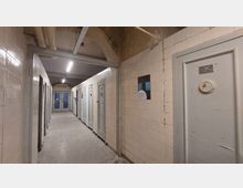 Ein schmaler, einfacher Flur mit abgenutzten weißen Fliesen an den Wänden und grauen Türen, die einzeln nummeriert sind. Im Hintergrund sind zwei doppelflügelige Türen mit Milchglasfenstern zu sehen, und die Decke zeigt sichtbare Rohre und Gebrauchsspuren.