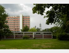 Flache Rasenfläche mit Bäumen im Vordergrund, dahinter ein einstöckiges Backsteingebäude mit der Aufschrift „Zum Abendmahl“ rechts an der Fassade. Im Hintergrund ein mehrstöckiges Wohngebäude und ein Kirchturm mit Kreuz.