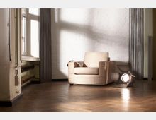 Ein heller Raum mit weißer Ziegelwand, Holzboden und großen Fenstern auf der linken Seite. Im Vordergrund steht ein beigefarbener Sessel, daneben liegt ein Studioscheinwerfer auf dem Boden.