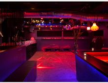 Ein moderner Clubraum mit roter und blauer Beleuchtung, einer zentralen Diskokugel, einer langen Bar und Sitzplätzen entlang der Wände. Der Raum hat eine industrielle Decke mit sichtbaren Lüftungsrohren und dekorativen Lampen.