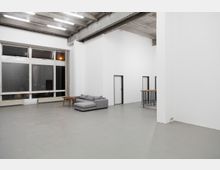 Industrieähnlicher Innenraum mit hohen Decken und sichtbaren Betonträgern, großen Fenstern auf der linken Seite, einer grauen Eckcouch mit Couchtischen und minimalem Mobiliar. Rechts befindet sich ein Geländer über einem Treppenaufgang sowie mehrere weiße Wände mit schlichten Türen.