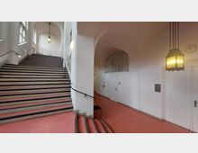 Breiter Treppenaufgang in einem historischen Gebäude mit weißen Wänden, hohen Decken und Rundbögen. Auf der rechten Seite des Raumes befinden sich weiße Türen und eine große, bogenförmige Fensteröffnung; Hängeleuchten und eine Wanduhr ergänzen die Einrichtung.