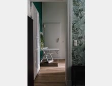 Schmaler Raum mit Holzboden, einer weißen Tür im Hintergrund und einem schlichten weißen Klapptisch mit passendem Stuhl. Die Wände sind teils grün, teils mit tropischem Tapetenmuster verziert, und es steht eine kleine Pflanze auf dem Tisch.