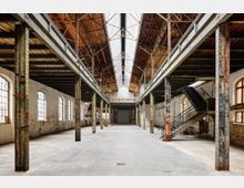 Innenraum einer alten Industriehalle mit hohen Decken, sichtbarer Stahlkonstruktion und einem Oberlicht aus Glas. Links und rechts befinden sich Reihen von großen, bogenförmigen Fenstern, während eine Metalltreppe auf der rechten Seite zu einer Galerieebene führt.