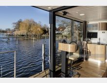 Innenraum eines Hausbootes mit modernem Holzinterieur, bestehend aus einer kleinen Küchenzeile, einem Steuerrad und einem Barhocker. Große Glasfenster bieten den Blick auf einen ruhigen See mit Stegen, Schilf und Häusern am Ufer.