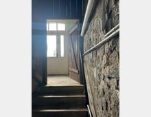 Steintreppe, die zu einer geöffneten Holztür mit Glasoberflächen führt; rechts eine grobe Steinmauer mit sichtbaren Kabeln, links Holzverkleidung. Durch die Tür fällt Tageslicht in die dunklere Umgebung des Treppenhauses.