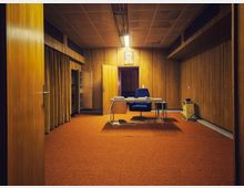 Innenraum eines retro wirkenden Büros mit holzvertäfelten Wänden und orangefarbenem Teppichboden, in der Mitte ein Schreibtisch mit Bürostuhl und sichtbaren Kabeln. Abgehängte Decke mit Leuchtstofflampe, seitlich ein Vorhang und im Hintergrund eine offene Tür sowie ein gerahmtes Porträt über der Tür.