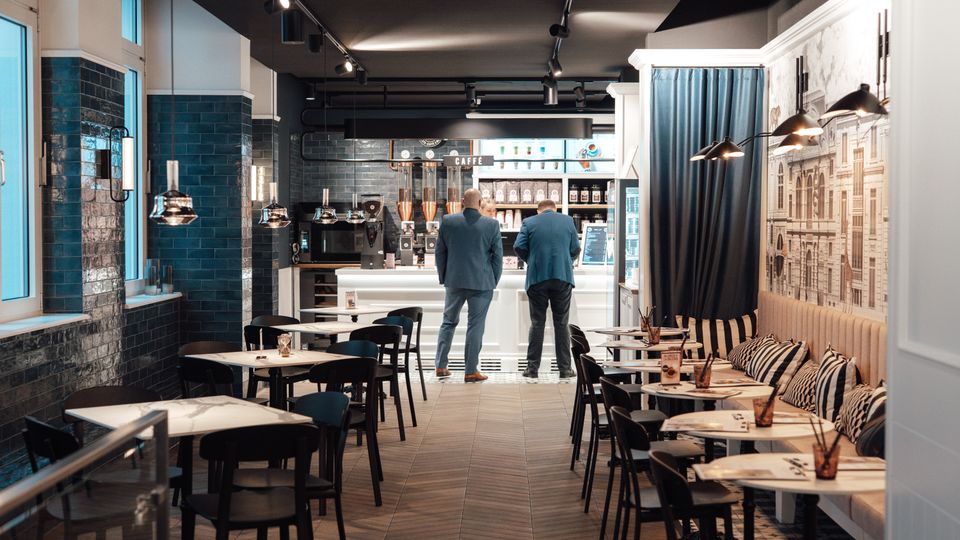 Ein modernes Café mit einer Mischung aus Sitzgelegenheiten an kleinen Tischen und einer gepolsterten Sitzbank an der Wand; die Rückwand ist mit einer großen Wandgrafik verziert, und die Bar im Hintergrund ist mit Kaffeemaschinen und Regalen ausgestattet. Schwarze Fliesen und dezente Hängeleuchten prägen die stilvolle Atmosphäre.