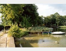 Kleiner See mit seichtem Wasser, umgeben von Bäumen und Schilf. Am Ufer stehen ein Holzsteg und ein kleines Boot, während einige Enten im Wasser schwimmen.
