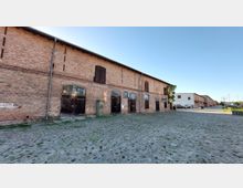 Backsteinlagerhaus mit großen Holztoren und hohen Fenstern auf einer gepflasterten Fläche. Im Hintergrund sind weitere ähnliche Gebäude sowie parkende Autos sichtbar, neben einer freien, sonnigen Umgebung.