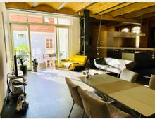Helles, modernes Loft-Wohnzimmer mit hohen Decken aus gelbem Klinker, großen Fenstern und einem Balkon. Im Vordergrund ein Esstisch mit Stühlen, im Hintergrund eine offene Küche, eine gelbe Liege und Pflanzen in Töpfen.