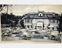 Historisches Foto eines Cafés mit parkähnlichem Außenbereich, zahlreichen Tischen und Stühlen, die um ein großes, mehrstöckiges Gebäude mit klassischem Walmdach und Fensterläden verteilt sind. Im Hintergrund sind Bäume und bewaldete Gegend zu sehen, während Sonnenschirme und Tischdecken auf eine gastronomische Nutzung hinweisen.