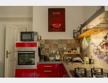 Compact kitchen with red lower cabinets, a built-in oven and microwave, and a long countertop with a sink and gas stovetop. Patterned tile backsplash, open shelves with bottles and utensils, and a poster on the wall above the counter.