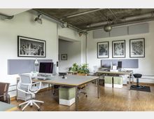 Großes, modern eingerichtetes Büro mit mehreren Arbeitsplätzen, ausgestattet mit Computern und Bürostühlen. Der Raum hat eine helle Wandgestaltung, Holzboden, eine Betondecke mit sichtbaren Rohrleitungen, mehrere gerahmte Schwarz-Weiß-Bilder an den Wänden sowie einige Zimmerpflanzen.