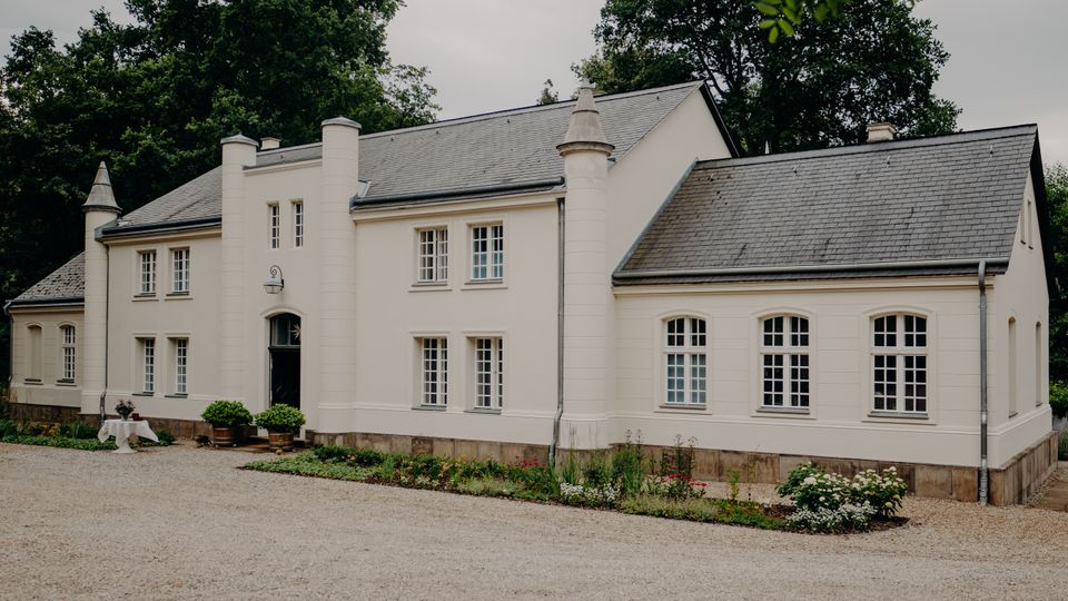 Weiße, zweigeschossige Villa im Landhausstil mit grauem Schieferdach, symmetrischer Fassade und zwei runden Türmchen am Eingangsbereich. Davor liegen ein Kieshof und ein schmaler bepflanzter Gartenstreifen, umgeben von hohen Bäumen.