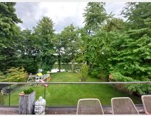 Großer, grüner Garten mit Rasenfläche und Spielgeräten wie Schaukel und Rutsche, umgeben von hohen Bäumen. Im Vordergrund eine Terrasse mit Glasgeländer, Sitzmöbeln und dekorativen Elementen, im Hintergrund ein kleiner See, teilweise verdeckt von den Bäumen.