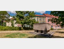Ein kleiner Stadtpark mit einer steinernen Gedenktafel, auf der Schriftzüge vermerkt sind, im Vordergrund. Im Hintergrund stehen mehrstöckige, farbige Wohnhäuser mit rotem Ziegeldach, umgeben von Bäumen und Sträuchern.