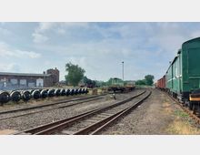 Gleisanlage eines alten Bahnbetriebswerks mit abgestellten Lokomotiven und Waggons, umgeben von Schienen, Ausrüstungsgegenständen und einem Backsteingebäude im Hintergrund. Links sind aufgereihte Zugräder und Garagentore des Werkstattgebäudes zu sehen.