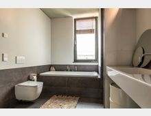 Modernes Badezimmer mit Wandtoilette, einer rechteckigen Badewanne und einem großen Fenster mit Holzjalousien. Der Raum ist in neutralen Farben gehalten, mit dunklen Fliesen und einer hellen Waschtischfläche mit rundem Spiegel.