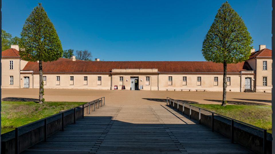 View from a shaded gateway down a wooden bridge with railings toward a long, pale courtyard building with a red tile roof and a central entrance. Two tall trimmed trees stand symmetrically in front, with open gravel ground and grass borders under a clear blue sky.