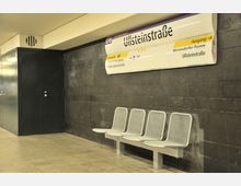 U-Bahn-Station mit dunkler Wandverkleidung, einer Sitzbank aus Metall mit vier Plätzen und einem Schild mit der Aufschrift "Ullsteinstraße" oben an der Wand.