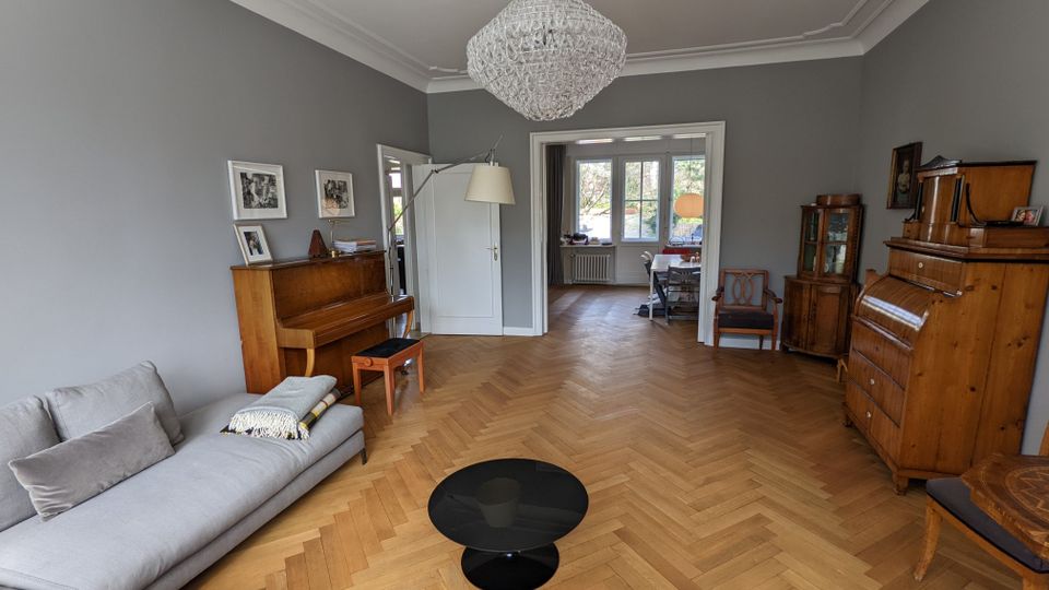 Großes Wohnzimmer mit grauen Wänden, Fischgrätparkett und einem Kristall-Kronleuchter an der Decke. Links stehen ein Sofa und ein Klavier, rechts mehrere Holzmöbel; durch eine breite Türöffnung führt der Blick in einen hellen Nebenraum mit großen Fenstern.