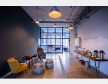 Offener Büro- und Aufenthaltsbereich mit modernen Möbeln, darunter Sessel, Hocker und ein Beistelltisch auf der linken Seite. Rechts steht ein langer Tisch mit Getränken und Snacks, im Hintergrund ist ein Besprechungsraum mit gläserner Trennwand zu sehen.