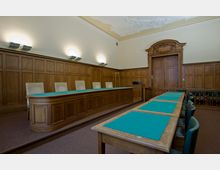 Holzgetäfelter Sitzungssaal mit einer erhöhten Richterbank und mehreren Stühlen, Tischen mit grüner Filzauflage und verzierten Deckenstuckelementen. Die Tür im Hintergrund ist aus dunklem Holz mit dekorativem Rahmen.