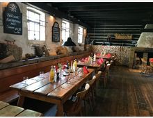 Rustikaler Gastraum mit Holztischen und -stühlen, gedeckt mit Gläsern, Getränken und roten Servietten. Im Hintergrund eine Wand aus gestapeltem Holz, Schiefertafeln mit Angeboten und Dekorationselemente wie Heuballen sowie ein offener Kamin.