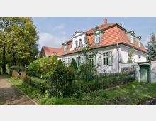 Großes historisches Wohnhaus mit weißer Fassade, grünen Fensterläden und rotem Walmdach. Das Gebäude ist von einem Metallzaun und dichtem Gartenbewuchs umgeben, und rechts führt eine grüne Tür durch eine gemauerte Gartenmauer.