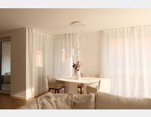 Esszimmer in einer modernen Wohnung mit einem weißen Esstisch und vier beigefarbenen Stühlen. Der Raum ist hell durch große Fenster mit weißen, durchsichtigen Vorhängen, ein minimalistischer weißer Kronleuchter hängt von der Decke, und auf dem Tisch steht eine Vase mit bunten Blumen.