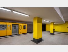Ein U-Bahnhof mit gelben Wänden und Säulen, beleuchtet durch langgestreckte Deckenlampen. Ein gelber U-Bahn-Waggon mit offenen Türen steht auf der linken Seite.