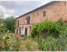 Backsteingebäude mit verblassenden roten Steinen, schwarzen hölzernen Fensterläden und bogenförmigen Fenstern, umgeben von wildem Bewuchs und Pflanzen, einschließlich Rosen und Kletterpflanzen. Der Himmel ist bewölkt, und die Umgebung wirkt ländlich und überwuchert.