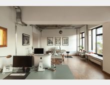 Helles, modernes Büro mit weißen Wänden, großen Fenstern und Holzfußboden. Schreibtische mit Computern und Büromaterialien stehen im Vordergrund, während an der Wand im Hintergrund gerahmte Bilder hängen.