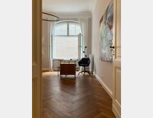 Ein helles Zimmer mit Fischgrätenparkettboden, einem großen, halbkreisförmigen Fenster mit weißen Vorhängen und einem Schreibtisch samt schwarzem Bürostuhl. An der rechten Wand hängt ein abstraktes Gemälde, und im Hintergrund ist eine hell gemauerte Wand durch das Fenster sichtbar.