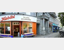 Eine Straßenansicht mit mehreren Geschäften, darunter ein Imbiss mit der Beschriftung „Habibi Falafel Schawarma“ in roten und goldenen Buchstaben auf einem weißen Schild. Rechts davon befinden sich ein historisches Gebäude mit Säulen und weitere Essenslokale, darunter ein Sushi-Restaurant. Fahrräder, Sitzbänke, und einige Bäume säumen die Gehwegkante.