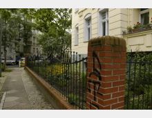 Ein Blick auf einen Bürgersteig neben einem eingezäunten Vorgarten mit Blumen und Sträuchern, flankiert von einem Ziegelpfeiler mit Graffiti. Im Hintergrund stehen mehrstöckige Wohnhäuser mit hellen Fassaden und großen Fenstern.