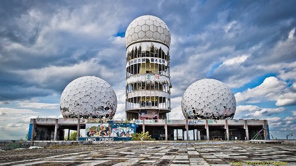 Verlassenes Radom-Gelände mit drei großen weißen Kugelkuppeln und einem zentralen zylindrischen Turm, dessen Fenster teils zerstört sind. Davor liegt eine weitläufige, rissige Betonfläche, die Gebäude sind mit Graffiti bedeckt, im Hintergrund ein wolkiger Himmel.