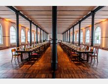 Großer Veranstaltungsraum in einem historischen Gebäude mit hohen Decken und schwarzen Eisensäulen; lange gedeckte Tafeln mit Stühlen stehen parallel zueinander auf einem Parkettboden, und große Bogenfenster lassen Tageslicht herein.